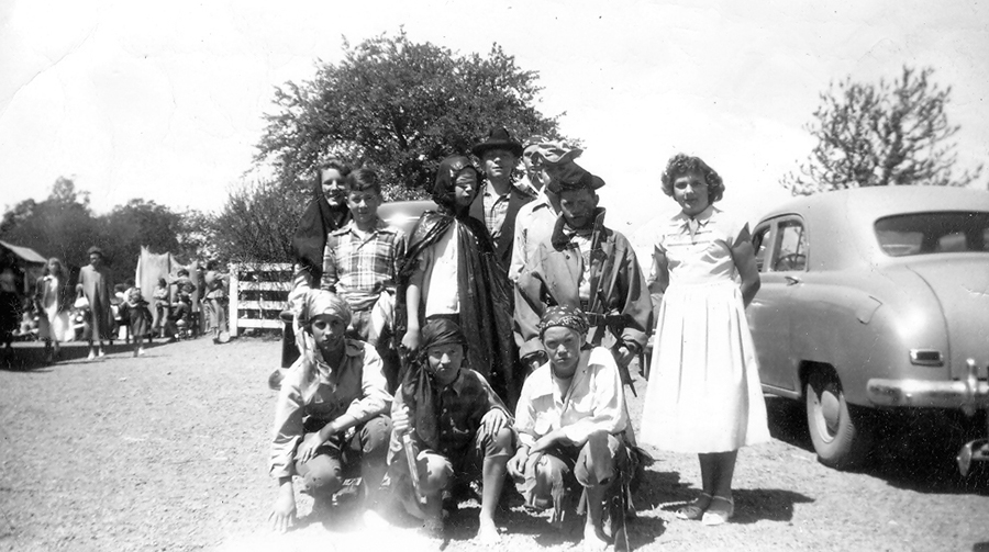 Black and white photograph of a group of approximately eleven students in costume. They appear to be dressed as pirates. There is a girl wearing a long white dress, so it may be a production of Peter Pan. The students are posed in the parking lot behind the school. A white fence and car are visible behind them. On the far left, parents and small children can be seen in the far distance walking toward the school.