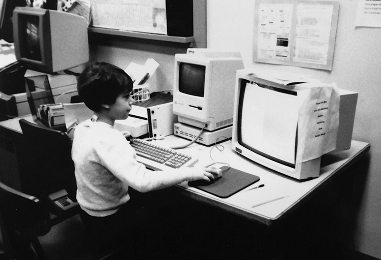 Black and white photograph of a student using a computer work station. The computer is a 1980s era Apple Macintosh. 