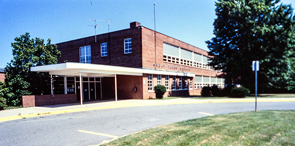 Color 35 millimeter slide photograph showing of the front exterior of Great Falls Elementary School. The picture is undated, but is believed to have been taken in the early 1980s. The building is a two-story brick structure with classrooms on both sides of the building separated by central hallways. The main office is located on the first floor near the main entrance. The main entrance doors are covered by a metal awning. A circular driveway curves in front of the building. In the center of the circle is a grassy area. The trees that were planted around the building in the 1950s have grown quite large.