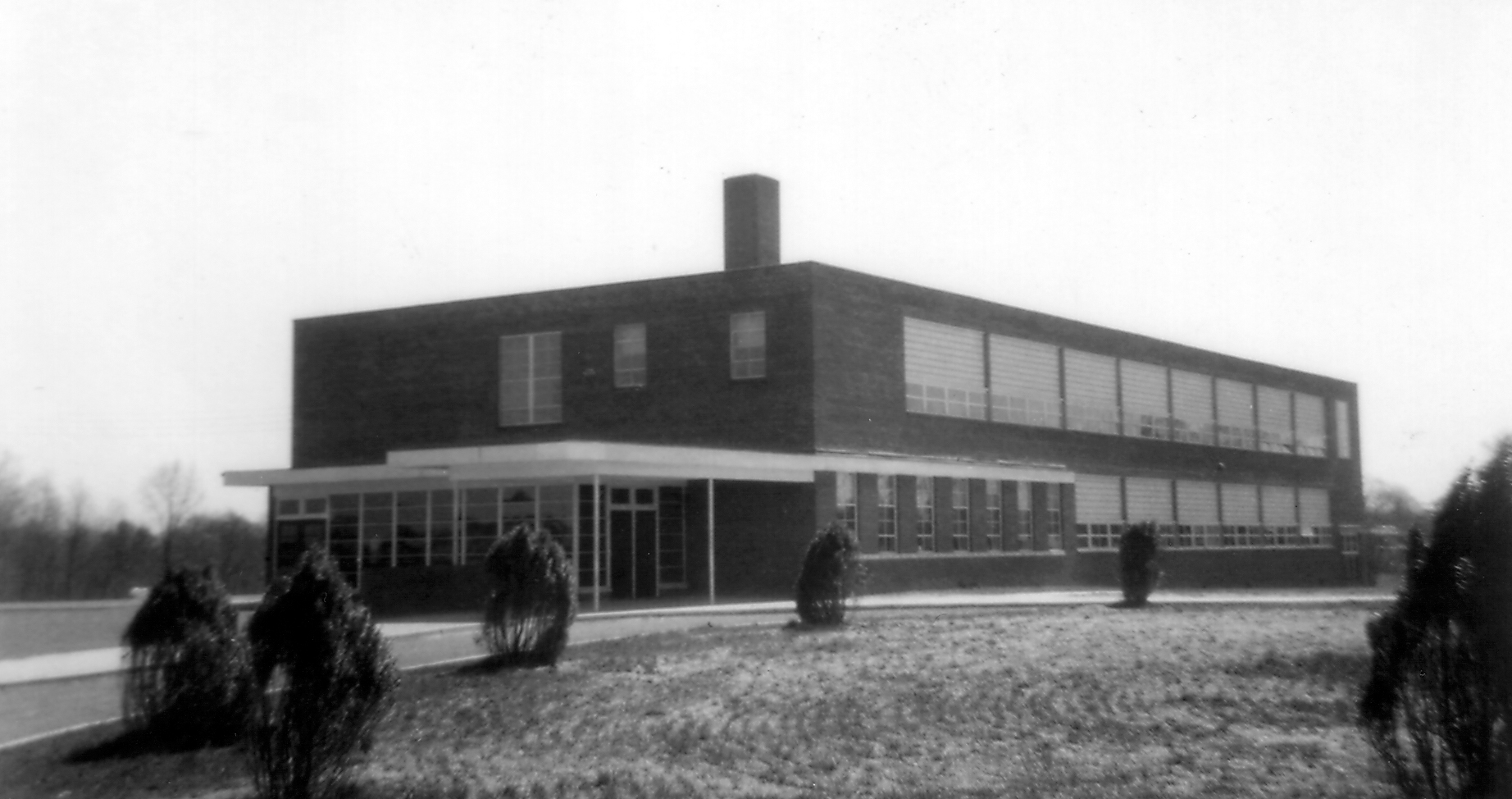 Black and white photograph of the front exterior of the new Forestville Elementary School taken in 1954. The building is a two-story brick structure with classrooms on both sides of the building separated by central hallways. The main office is located on the first floor near the main entrance. The main entrance, a set of double doors surrounded by windows, is covered by a metal awning. A circular driveway curves in front of the building. In the center of the circle is a grassy area where several trees or tall shrubs have been planted. 