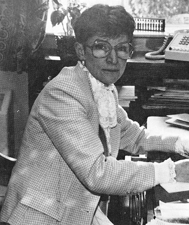Black and white photograph of Principal Marie Canny from Great Falls Elementary School’s 1986 to 1987 yearbook. She is sitting at her desk looking up from her paperwork.