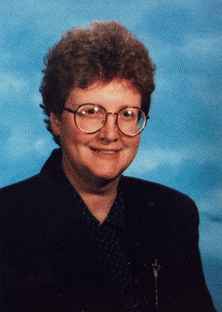 Head and shoulders portrait of Principal Elizabeth Henderson from Great Falls Elementary School’s 1994 to 1995 yearbook. She has glasses and is wearing a dark colored jacket, and is posed in front of a blue background.