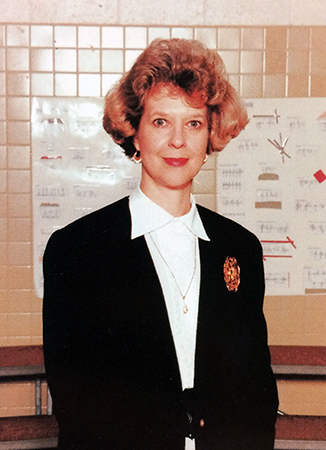 Head and shoulders portrait of Principal Helen Davis from Great Falls Elementary School’s 1996 to 1997 yearbook. She is standing in the cafeteria, and is wearing a white blouse with a dark jacket that has a brooch pinned to the lapel. 
