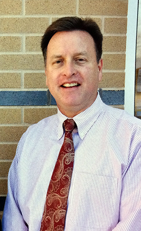 Color head and shoulders portrait of Principal Leighty from Great Falls Elementary School’s 2011 to 2012 yearbook. He is wearing a blue and white striped shirt with a red paisley tie. He is standing outside the school next to a brick wall.