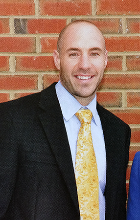 Color head and shoulders portrait of Principal Lonnett from Great Falls Elementary School’s 2012 to 2013 yearbook. He is wearing a blue shirt with a yellow tie and a black blazer. He is standing outside the school next to a brick wall.