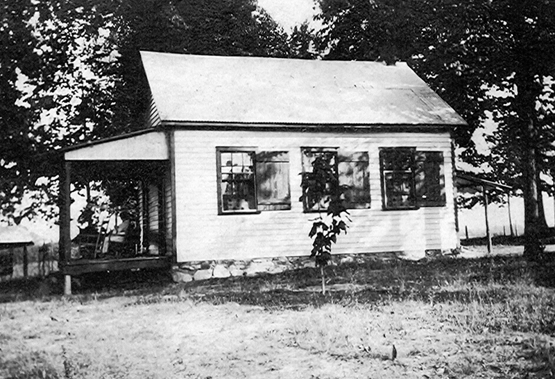 Black and white photograph of the Forestville Colored School after it had been converted into a dwelling. The building is a small, single story structure with a tin roof and white clapboard siding. There are three windows visible on the side of the building with wooden shutters. A person can be seen sitting in a rocking chair in the shade on the front porch. The building has a stone foundation and is surrounded by trees. 