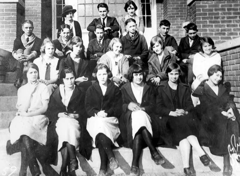 Black and white photograph of Forestville High School students. The students are seated on the steps leading to the main entrance of the school. 17 students and three adults are pictured. Most of the students are wearing winter coats. The girls are seated in the first two rows, the boys in the third row, and the adults in the back row. 