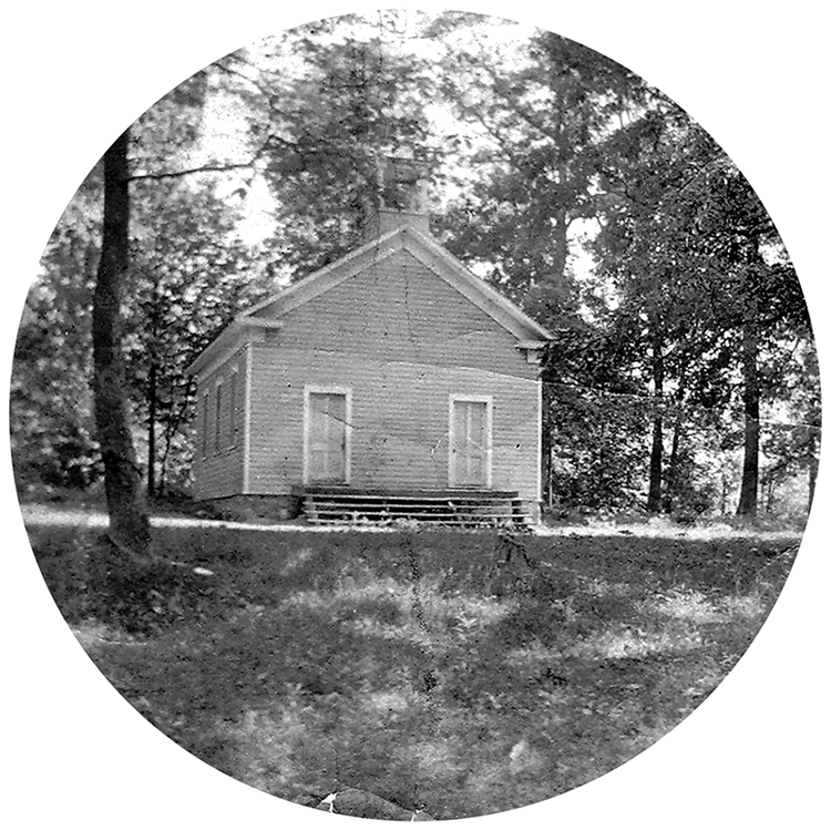 Black and white photograph of the Forestville one-room schoolhouse taken in 1899. The building is a one-story structure with a pitched roof and a bell tower. The building has three windows on the side facing the camera, and two doors on the front of the building facing Georgetown Pike. The school is surrounded by trees.