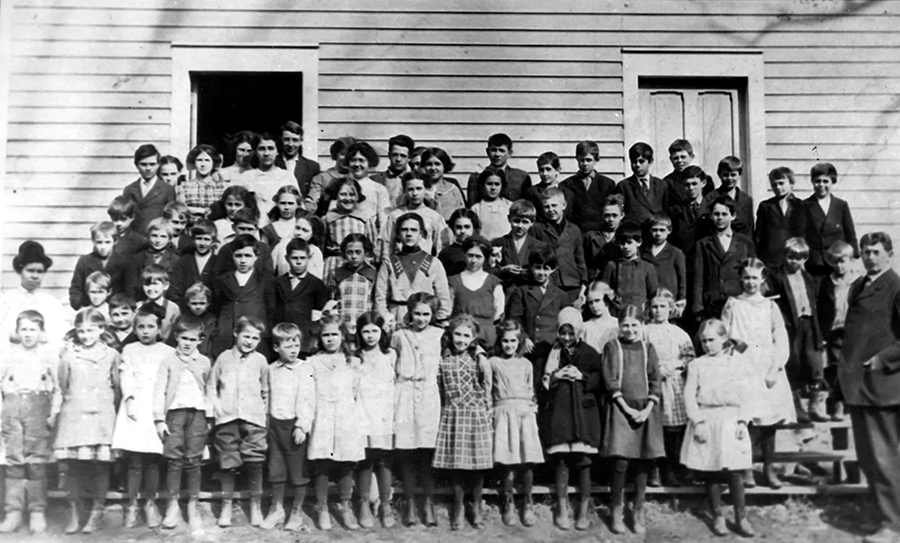 Black and white class photograph of the Forestville School taken in 1913. Two teachers and approximately 72 children are pictured. The children range between six and eighteen years in age. They are standing in rows on the wooden steps in front of the schoolhouse. The school has clapboard wood siding and two doors. The door on the left is open.