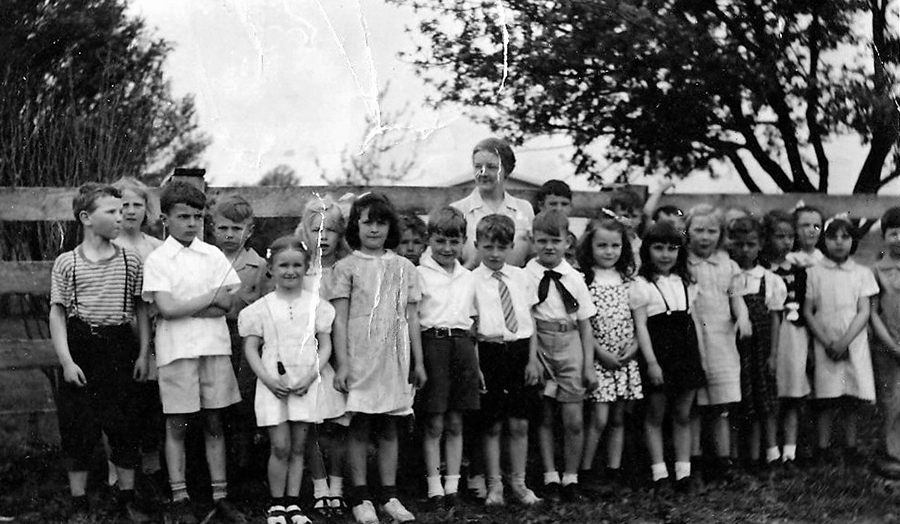 Black and white photograph of Forestville Elementary School’s first grade class in 1942. Approximately 24 students and their teacher are shown. They are standing next to a fence outside the school with trees in the background.