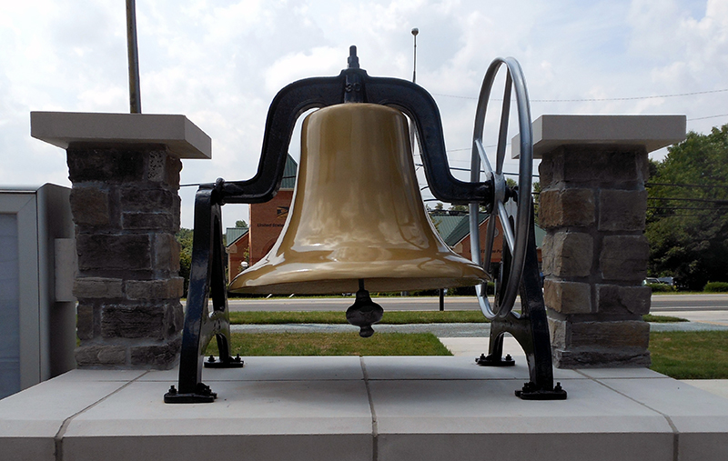 Photograph of the bell from the one-room schoolhouse at Forestville. It was also used from approximately 1922 to 1949 on top of the Forestville high school and elementary school building. The bell is painted gold and is mounted in a black-painted steel housing. There is a wheel to the right of the bell on which a cord would have been wound so the bell could be rung. The bell and housing have been mounted to a brick pedestal outside the fire station.