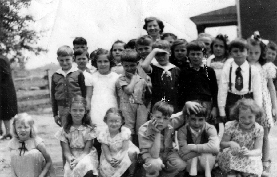 Black and white photograph students and their teacher standing together outside behind Forestville Elementary School. The rear entrance to the building is visible in the distance behind them. Two female adults and approximately 27 children are pictured. One adult is standing behind the students and the other is on the left. She is partially obscured because she is walking out of the picture. 