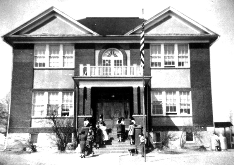 Black and white photograph of the front exterior of the new Forestville School. It is a two-story structure constructed of brick and has a basement. The main entrance to the building is a set of double doors at the top of a staircase. A porch awning, supported by brick pillars, is over the steps. The top of the porch has a balcony with a white wooden railing. Behind it, a set of double doors or windows is visible on the second floor. The windows have a glass archway above them. Four sets of windows are visible on the front of the building, one on each side of the entrance and similar ones above on the second floor. There is a flag pole out front and the United States flag is visible at the top. People can be seen milling about on the steps in front of the building.