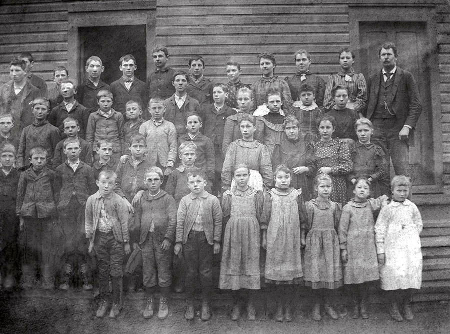 Black and white class portrait from the 1888 to 1889 school year at the Oak Grove School in Forestville. The students are arranged in four rows on the steps in front of the schoolhouse. The building has wooden clapboard siding and two doors, one of which is propped open. 42 children and their teacher are pictured. The children range between 6 and 18 years of age. The youngest children are standing on the bottom rows, and the group is evenly divided with the girls on the right and the boys on the left. 
