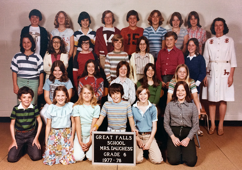 Color photograph from the 1977 to 1978 school year showing Mrs. Dauchess’ sixth grade class. 26 children and their teacher are pictured. The children are arranged in four rows along the wall in the gymnasium. Mrs. Dauchess is standing in the back row on the far right.