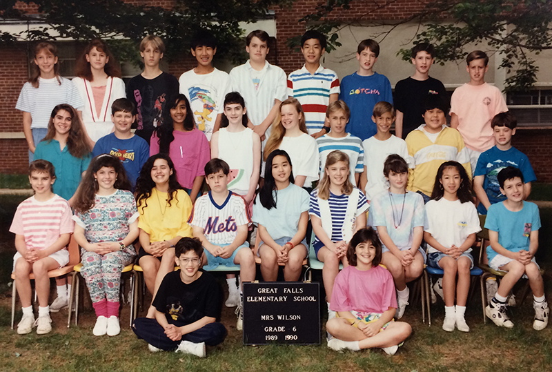 Sixth grade class photograph taken during the 1989 to 1990 school year. 29 children are pictured. They are arranged in three rows outside the building. The first row is seated on chairs, the second row is standing, and the third row is standing on chairs. The children are underneath the tall trees that lined the front of the original classroom wing of the building.