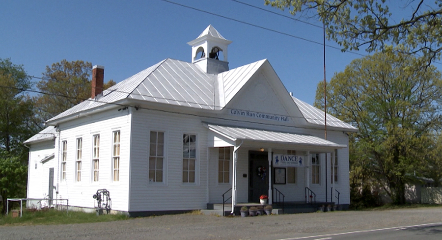 Color photograph of the Colvin Run Community Hall in 2013. The building is an old schoolhouse. It still retains the bell tower and bell on top of the building. The school had two classrooms and an auditorium. It was built on the south side of Colvin Run Road west of its intersection with Walker Road, on the site of an earlier schoolhouse in the village that burned down. The building faces Colvin Run Road. It has white clapboard siding and a gray tin roof. There are tall windows on all sides and a red brick chimney juts out from the roof above the classroom on the left. Today the building is used as a dance hall. 
