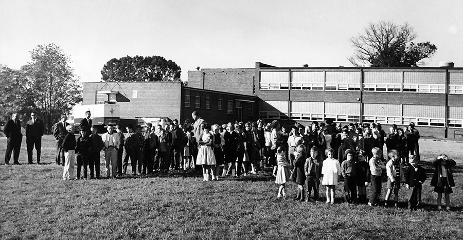 Black and white photograph of Great Falls Elementary School taken in 1956. The student body is standing outside the school on the field behind the building. The students are all wearing coats, but some of the trees in the distance still have leaves so it could be a crisp fall day. Several adults can be seen walking among the students and watching from a distance. It is unclear what the students are doing. It may be a fire drill or something similar because some of children appear to be lined up in rows.