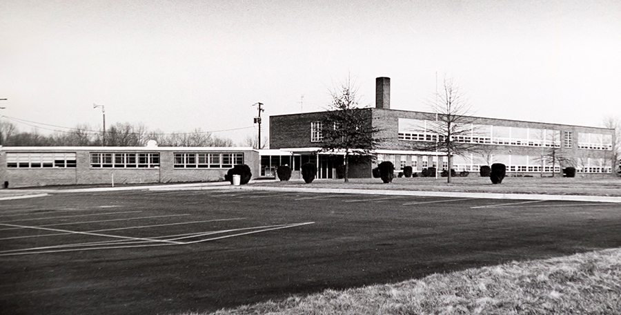 Black and white photograph of the front exterior of the new Forestville Elementary School taken circa 1969. The original section of the building is a two-story brick structure with classrooms on both sides of the building separated by central hallways. The main office is located on the first floor near the main entrance. The main entrance, a set of double doors surrounded by windows, is covered by a metal awning. A single-story addition has been constructed to the left of the main entrance. The circular driveway has been removed and the area is now a parking lot.