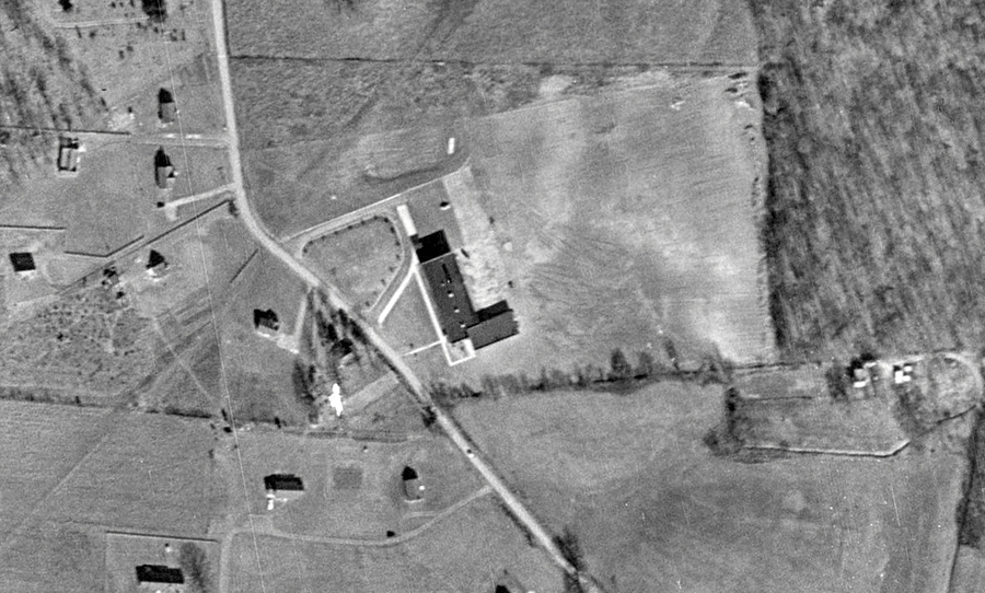 Black and white aerial photograph of Great Falls Elementary School taken in 1954. The L-shaped building is seen from directly overhead. The countryside around the school is largely farm fields and forests. There are a few houses scattered on large lots on the west side of Walker Road across from the school. 