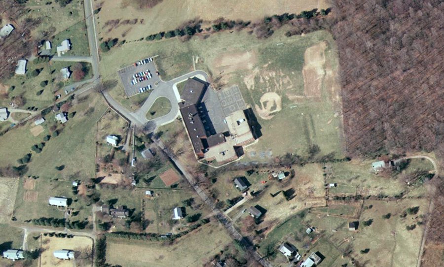 Color aerial photograph of Great Falls Elementary School taken in 1976. The building is seen from directly overhead. The countryside around the school still has some farm fields and forests, but many of the large lots have been subdivided and new homes have been built. The building, originally shaped like the letter L, more closely resembles the letter C in shape now. The new addition has almost doubled the size of the original building. The brick walls and rooftop of the addition are a different color from that of the original building. The roof of the new wing is light beige, the older roof is dark grey in color.