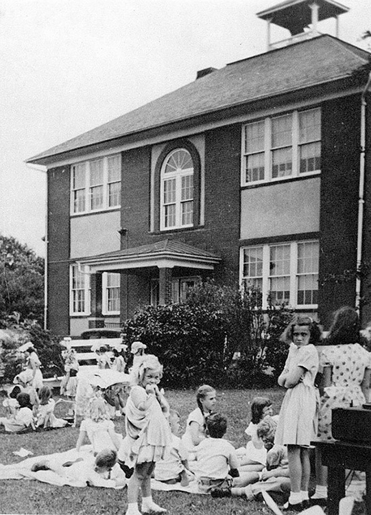 Black and white photograph students playing behind Forestville Elementary School. Mostly girls are shown, playing on the grassy lawn. 