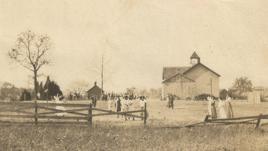 Sepia-toned photograph of the three-room school at Floris taken in 1910. The building is in the center of a large field surrounded by a wooden fence. Children can be seen playing outside in the field.