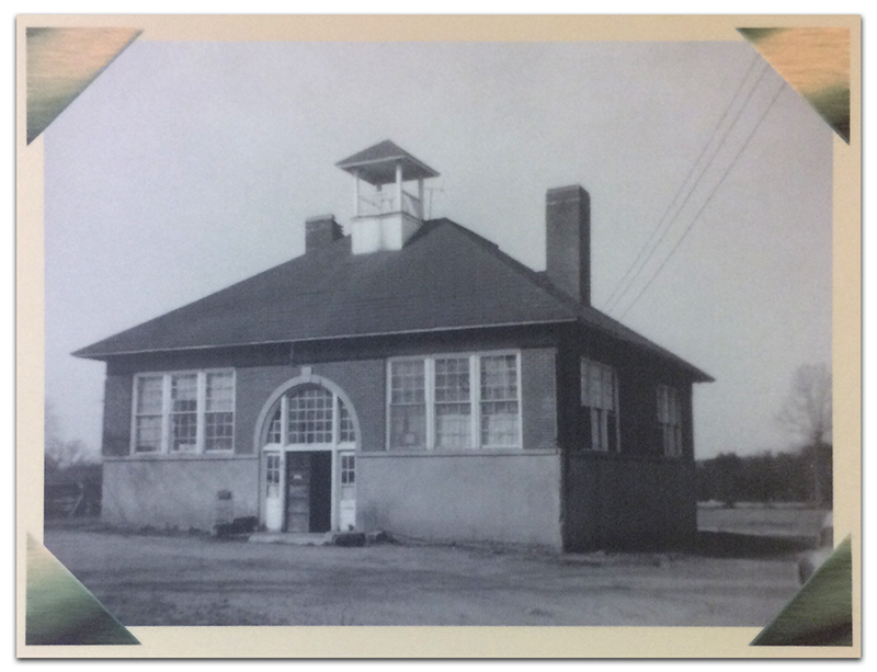 Black and white photograph of the Floris School constructed in 1911. The building is a single story structure with four classrooms and a central hallway. The school has two brick chimneys and a bell tower. The children enter the building through double wooden doors beneath a large arched window.