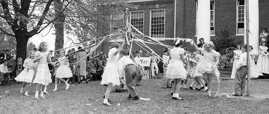 Black and white photograph of students performing a maypole dance in front of a school. A group of students are walking around the maypole, some clockwise, some counter clockwise. Each is holding a long piece of fabric. As they walk, the students alternate as they pass one another, forming a braid of sorts around a tall wooden pole in the center of the circle. The children are dressed in very nice clothing – the girls in white dresses and the boys in shirts and slacks. Parents and other students are seated off to the side watching the maypole dance.