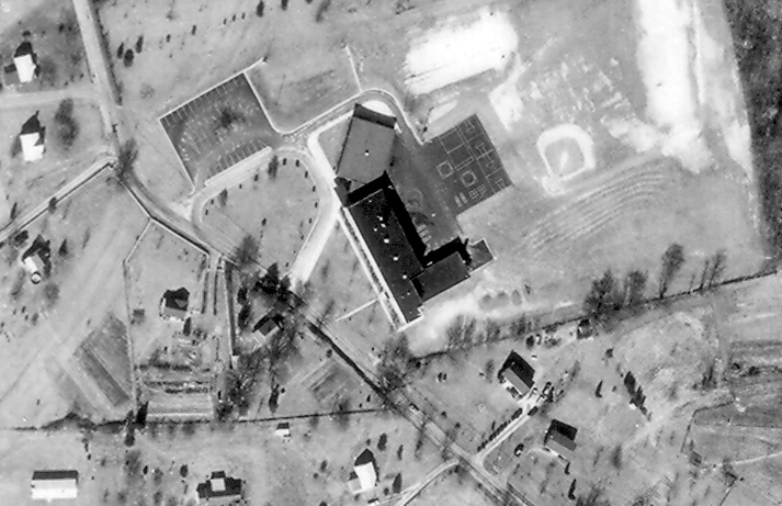 Black and white aerial photograph of Great Falls Elementary School taken in 1968. The building is seen from directly overhead. The countryside around the school is largely farm fields and forests. There are a few houses scattered on large lots on the west side of Walker Road across from the school. The photograph is an animated gif image with colored circles that appear over portions of the image. A blue circle highlights a two-story, two-classroom addition that was constructed in 1955. A red circle shows the six-classroom addition that was constructed on the north side of the building near the main entrance. A yellow circle highlights an old schoolhouse across Walker Road from Great Falls Elementary. Called the Forestville Colored School, you can learn more about this old schoolhouse on the 1870 to 1922 school history page.