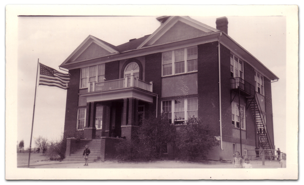 Black and white photograph of the front and right side exterior of Forestville Elementary School taken in 1942. It is a two-story structure constructed of brick and has a basement. The main entrance to the building is a set of double doors at the top of a staircase. A porch awning, supported by brick pillars, is over the steps. The top of the porch has a balcony with a white wooden railing. Behind it, a set of double doors or windows is visible on the second floor. The windows have a glass archway above them. Four sets of windows are visible on the front of the building, one on each side of the entrance and similar ones above on the second floor. There is an identical placement of four windows on the side of the school. A metal fire escape staircase leads down from the second floor to the ground on this side of the building. There is a flag pole out front and the United States flag is visible at the top. Several students can be seen outside the building.