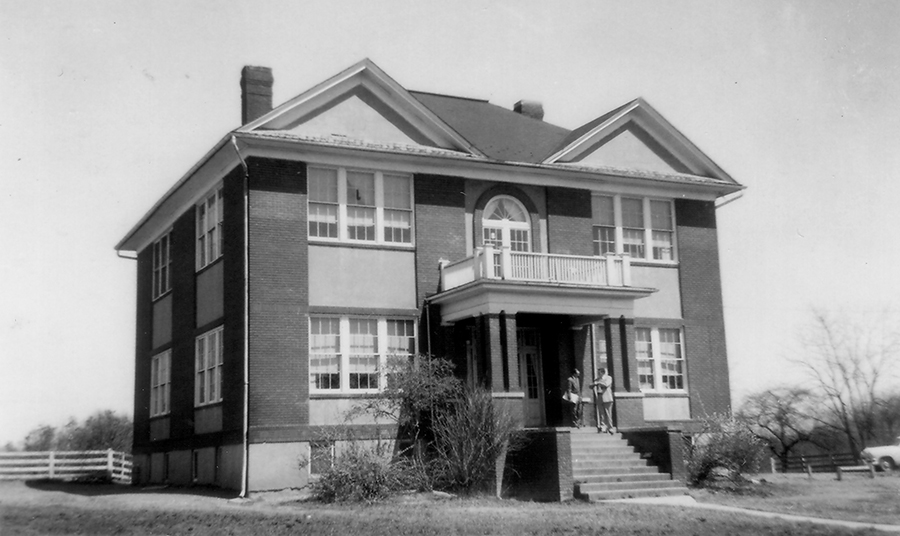 Black and white photograph of the front and left side exterior of the old Forestville Elementary School taken in 1954. It is a two-story structure constructed of brick and has a basement. The main entrance to the building is a set of double doors at the top of a staircase. A porch awning, supported by brick pillars, is over the steps. The top of the porch has a balcony with a white wooden railing. Behind it, a set of double doors or windows is visible on the second floor. The windows have a glass archway above them. Four sets of windows are visible on the front of the building, one on each side of the entrance and similar ones above on the second floor. There is an identical placement of four windows on the side of the school. The bell tower has been removed. Two men are standing at the top of the steps at the main entrance.