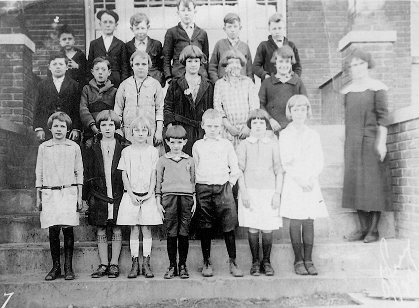 Black and white photograph of students at the new Forestville brick schoolhouse taken in 1924. 19 students and a female teacher are pictured. The children are arranged in three rows, standing on the steps in front of the building.
