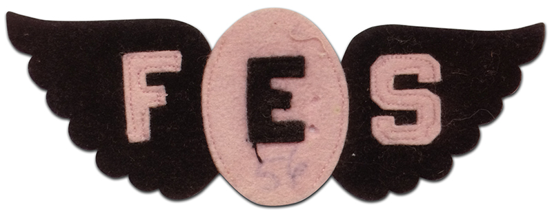 Photograph of a felt school spirit patch from Forestville Elementary School. The hand-made patch was created during the mid-1950s. It is approximately five inches across and features an oval at the center with wings on the left and right sides of the oval. The wings are made out of black felt and the oval is made out of pink felt. The letters F, E, and S, have been sewn on to the patch. The letter F, cut from pink felt, is sewn onto the left wing. The letter E, cut from black felt, is sewn onto the center of the oval. The letter S, cut from pink felt, is sewn onto the right wing. The letters are held in place with hand-stitched pink thread.