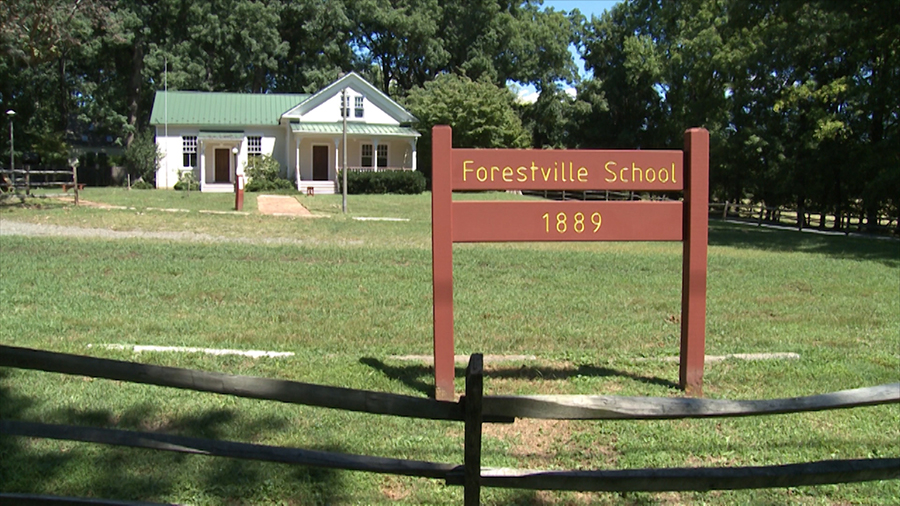 Color photograph of the old Forestville School taken in 2014. The building is seen from Georgetown Pike at the crest of a hill. A brown Fairfax County Park Authority sign with the words Forestville School, 1889, has been placed on the property very close to the roadway.