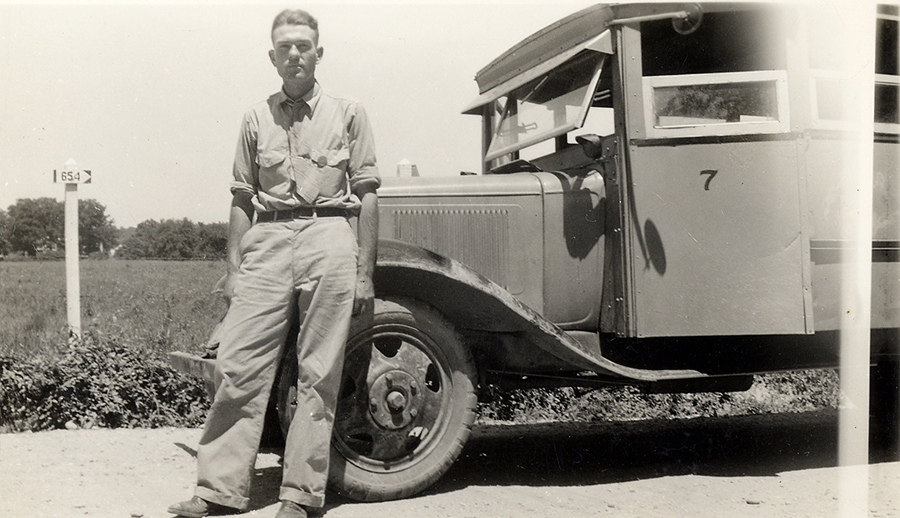 Black and white photograph of Fred Jones standing next to his school bus in 1941. Jones is leaning back against the engine. The side of the bus, showing the engine compartment, driver’s seat, and the first row of student seating is shown. The bus is parked on a dirt road next to a farm field.