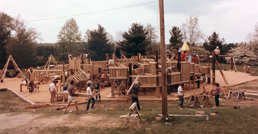 Photograph showing a wide view of the entire playground. The structures are made out of wood. The playground has swings and several different types of structures on which to climb and play. Adults can be seen working on various parts of the playground.