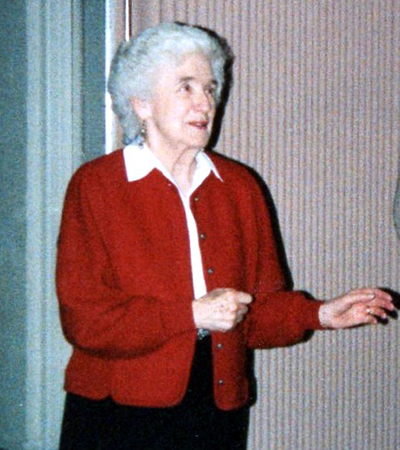 Color photograph of Principal Winston taken several years after her retirement. She has white hair and is wearing a white blouse and red sweater. She appears to be in mid-conversation with someone to her left who is not pictured.