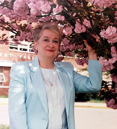 Color photograph of Principal Gina Ross taken in 1992. She is standing beneath one of the cherry trees in front of the school. The tree is in bloom with large clusters of delicate pink blossoms.