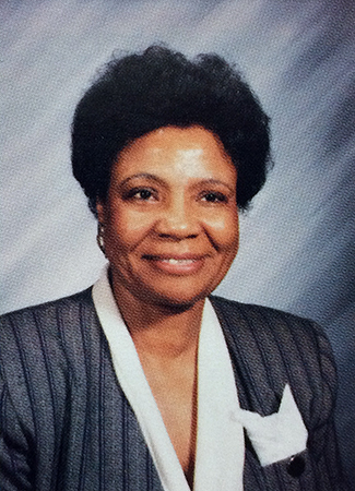 Head and shoulders portrait of Principal Dorothy Clark from Great Falls Elementary School’s 1994 to 1995 yearbook when she was an assistant principal. She is seated in front of a striped grey background and is wearing a white blouse and dark grey jacket with a white pocket handkerchief.
